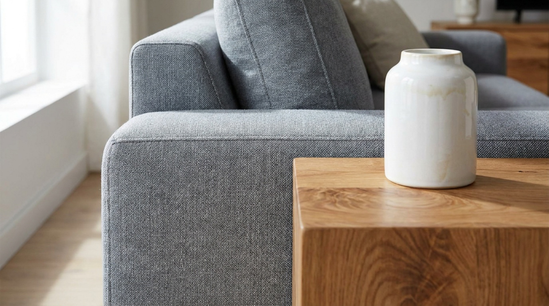Nahaufnahme einer grauen Stoffcouch neben einem Holztisch mit weißer Keramikvase.